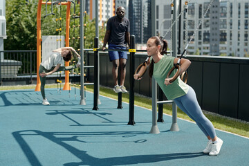 Young pretty and fit woman in activewear hanging on still rings on sports ground while doing...