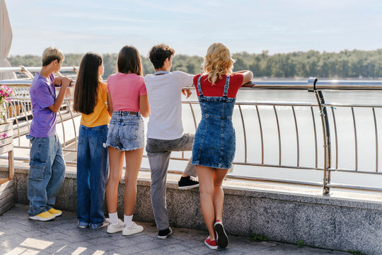 Back view, positive teenage boys and girls standing by urban street railing outdoors