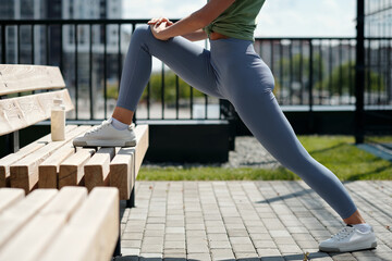Cropped shot of young fit woman in blue leggins and white sports shoes standing by bench anf doing stretching exercise for leg muscles