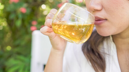 A woman in white is sipping a glass of hot tea in the morning with bokeh garden backgrounds. Concept for Herbal drinks, Alternative medicine, Healthy food, Relaxation, Leisure activity.