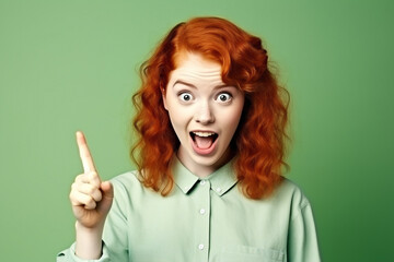 Excited young ginger teenager girl smiling and pointing index finger up at copy space expressing wow emotion standing isolated on green background studio copy space, advertisement concept. High