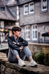 Young woman in black leather jacket with short dark hair sitting on stone wall in old german city