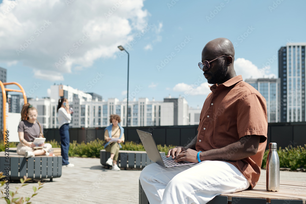 Poster young serious businessman typing on laptop keyboard while sitting on bench in urban environment on s