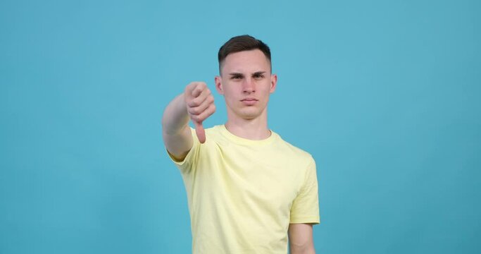 Displeased Caucasian man jn the blue background holds his hand up with his thumb pointing downward, showing the thumbs down gesture. His facial expression reflects dissatisfaction or disapproval.
