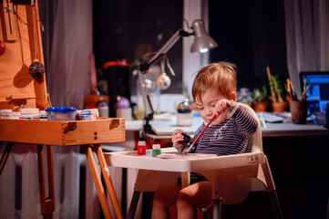 Lottle toddler boy sitting in chair for babies happy to play with brush and jars of paint at home