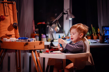 Lottle toddler boy sitting in chair for babies happy to play with brush and jars of paint at home