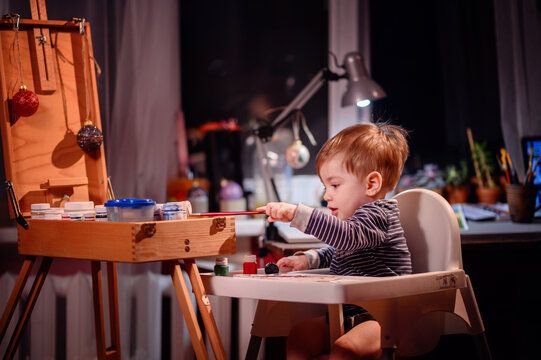 Lottle toddler boy sitting in chair for babies happy to play with brush and jars of paint at home