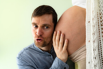 Curious father holds the naked baby bump of the expectant mother and listens to the baby. Green background. Bright shot.