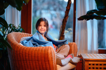 Little praschooler serious girl sits with big orange tea cup covered in blue plaid in straw armchair