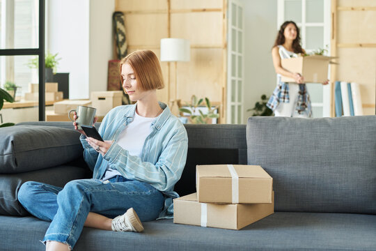 Young woman in denim casualwear sitting on soft comfortable couch in living room of new apartment, having tea and texting in smartphone