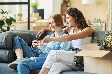 Happy young woman pointing at smartphone screen while watching online video with her girlfriend at home and discussing something curious