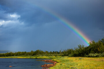 Rainbow by the side of the Lainio river in the Swedish Lapland with dramatic dark sky and clouds in the background on summer afternoon.