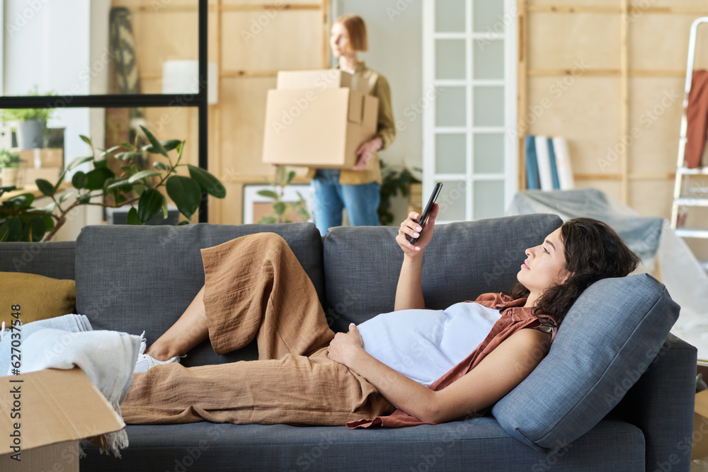 Wall mural young pregnant woman holding mobile phone in front of her face while relaxing on soft couch at home 