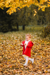 A little girl in a red warm coat is playing with yellow leaves, having fun and smiling sitting on a plaid plaid in an autumn park outdoors in fall. Selective focus