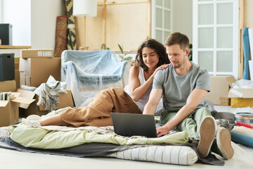 Young affectionate couple sitting on checkered plaid on the floor of living room and looking at laptop screen while man typing on keyboard