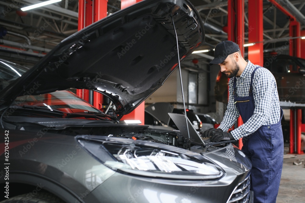 Poster A bearded auto-mechanic in working uniform is standing next to a broken car and using the special laptop for car maintenance. An auto-mechanic in workshop repairs car.