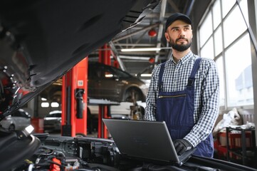 A bearded auto-mechanic in working uniform is standing next to a broken car and using the special laptop for car maintenance. An auto-mechanic in workshop repairs car.