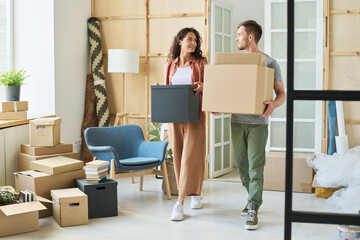 Young couple carrying packed boxes and looking at one another while moving along spacious living...