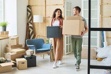 Young couple carrying packed boxes and looking at one another while moving along spacious living room of new apartment during relocation
