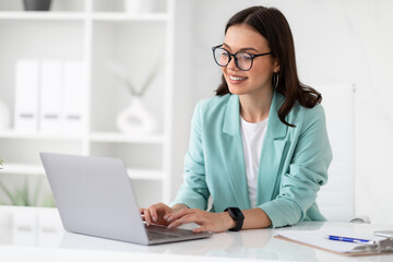 Cheerful millennial caucasian lady in suit and glasses typing on laptop at workplace