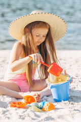 Cute child playing on the beach on summer holidays. Little girl in straw hat having fun with a sand...