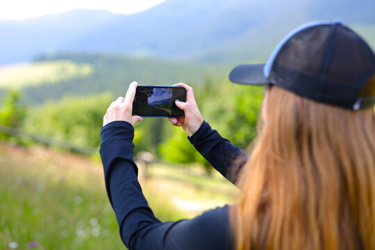 Woman taking a photo of a mountain landscape with their phone. The person is standing in a grassy field with wildflowers.