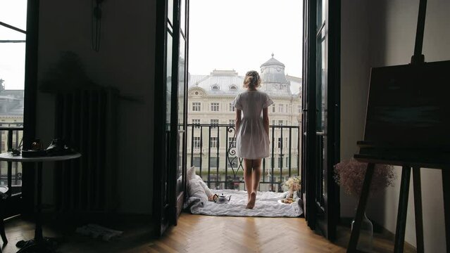 A Gyrl Grec The Morning On The Balcony Of A Hotel In A Big Chit. The Girl Gos Ut On The Balzoni And Looks Around The Chiti. Rer Is Blowing From The Apartment