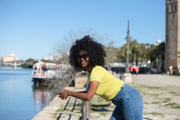 Fototapeta na wymiar Young and beautiful black woman with afro hair and sunglasses wearing jeans and yellow shirt leaning on the railing overlooking the river in seville.