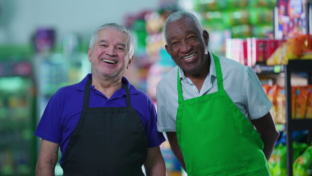 Happy diverse senior workers standing at grocery store smiling at camera wearing uniforms. Older Brazilian staff workers