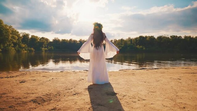Art portrait happy young woman walks by river bank water hand raised to blue sky sun light green forest trees back rear view girl enjoy summer nature. herbal wreath long hair white dress slow motion