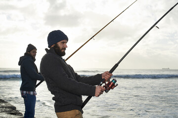 Secretly hoping hell hook the bigger fish...Shot of two young men fishing at the ocean in the early...