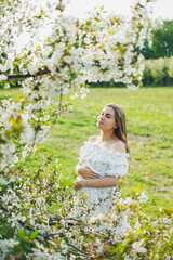Fototapeta na wymiar A young pregnant woman in a white dress walks in a blossoming apple orchard. Happy pregnant woman. Happy future mother
