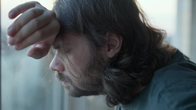 Depressed man staying alone near window with stressed problem face, closed eyes. Pensive, thoughtful. Denial, Bargaining emotions. Mental health, man depression, despair concept. Long hair, beard