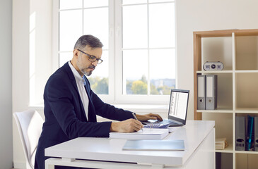 Male accountant or bookkeeper working in office. Mature man in suit and glasses sitting at desk, using modern laptop computer, working with business data, and taking notes in accounting book