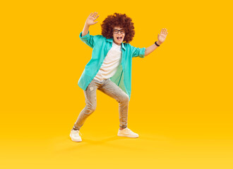 Fototapeta na wymiar Funny joyful child in casual clothes dancing and having fun in fashion studio. Happy little boy in curly wig, turquoise shirt, white T shirt and glasses dancing or waving hello on orange background
