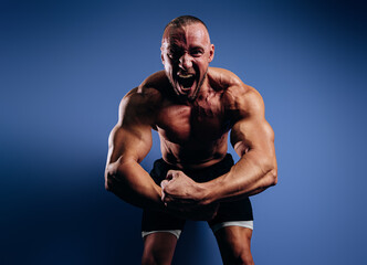 Super emotional bodybuilder screaming and showing muscles on blue background. Fit man with muscular...