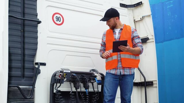 Professional truck driver checking using tablet computer and standing by long vehicle. Transportation service
