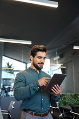 Smiling busy young Latin business man entrepreneur using tablet standing in office at work. Male...