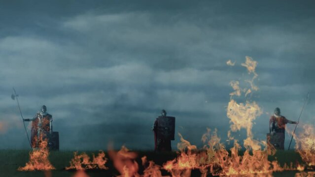 Three Roman soldiers declare victory on a battlefield engulfed in flames and heat on a cloudy day. Slow motion