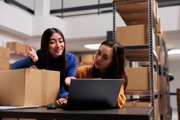 Storehouse supervisor explaining operator delivery tracking on laptop while working at desk in storage room. Warehouse women employees managing shipping operation on computer
