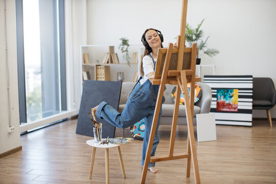 Excited lady in denim outfit creating art with paint and brush while moving to loud tracks in headphones. Enthusiastic female adult in high spirits expressing vision and mastery on canvas in workshop.