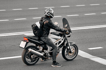 A man on a black and white road bike stands at a traffic light. A motorcyclist in a leather jacket is ready to start the journey. Biker made a stop on the road.