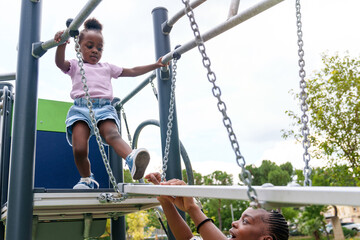 Fearful yet brave, a 5-year-old African girl cautiously steps on a wobbly bridge in a vibrant park,...