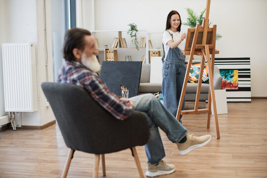 Attractive brunette lady in denim overalls sketching on canvas while looking at older man sitting in chair at workshop. Young caucasian artist depicting live model full body in art studio interior.