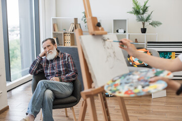 Selective focus of aged man in casual clothes sitting in chair behind wooden easel with painting in...