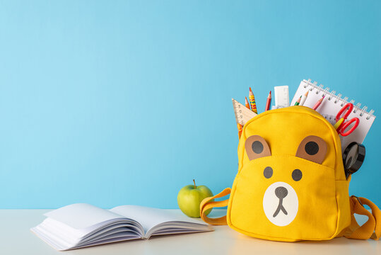 School organization. Side view photo of teddy-bear schoolbag filled with school essentials - pens, pencils, notebook, and more. Open book and an apple bring a playful element to the scene