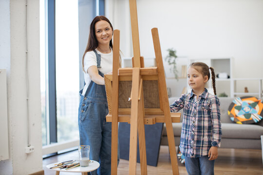 Portrait of beautiful female and little child in casual clothes standing behind easel with painting in workshop. Attentive art person teaching inquisitive small student mixing oil paints on paper.