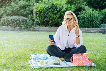 Middle aged caucasian woman sitting on green lawn, eating healthy lunch and using phone. Having...