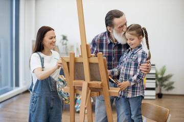 Loving aged grandfather in casual wear kissing little kid while smiling lady doing sketches on...