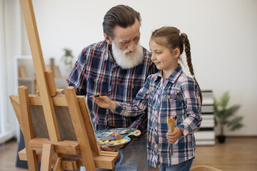 Close up view of senior grandfather and little child in similar outfits working on interesting art project. Caring grandpa teaching how to fill drawing in with color from palette in home studio.